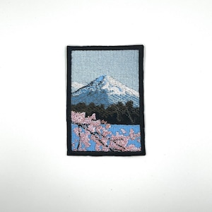 Könnte beinhalten: Gestickter Aufnäher mit einer Bergszene mit schneebedecktem Gipfel, grünen Bäumen und einem See im Vordergrund. Im unteren rechten Eck sind rosa Kirschblüten zu sehen.