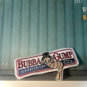 Könnte beinhalten: Weißer gestickter Aufnäher mit roten, blauen und goldenen Akzenten. Der Aufnäher zeigt den Text "Bubba Gump Shrimp Co." und eine Karikatur einer Garnele mit Hut.