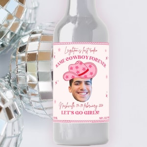 May include: A small clear glass bottle with a silver cap and a pink and white label. The label has a photo of a person wearing a pink cowboy hat and the text "Let's Go Girls!" and "Same Cowboy Forever".