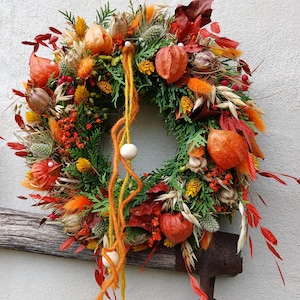 Könnte beinhalten: Ein runder Kranz aus grünen Zedernzweigen, orangefarbenen Lampionblumen, roten und gelben Trockenblumen sowie braunen Samenkapseln. Zwei lange orangefarbene Wollfäden hängen von oben herab, verziert mit drei glatten, runden Holzperlen.