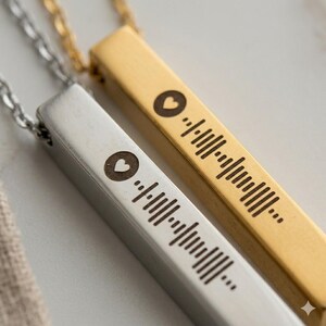 Peut inclure: Gros plan sur deux colliers pendentifs. L'un est doré, l'autre argenté. Chacun a une barre rectangulaire avec un symbole de cœur et une série de lignes, ressemblant à un code musical. Les colliers sont sur une surface blanche.