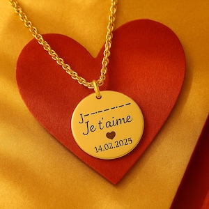 Puede incluir: Un collar de oro con un colgante redondo que dice "Je t'aime" y "14.02.2025". El colgante está sobre un fondo rojo en forma de corazón.