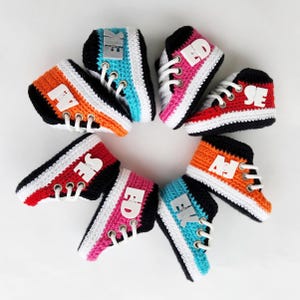 Peut inclure: Une collection de mini-baskets crochetées colorées disposées en cercle. Chaque chaussure présente une palette de couleurs différente, notamment orange, rouge, rose et bleu, avec des lacets blancs et des accents noirs. Des lettres blanches sont visibles sur les côtés.