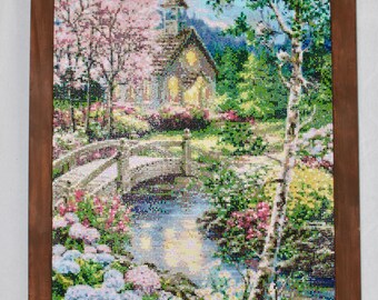 May include: A framed, detailed artwork depicting a serene landscape with a church, bridge, and flowing water. The scene is filled with colorful flowers, trees, and a soft, natural light. The frame is a dark brown color.