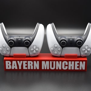 Può includere: Due controller di gioco wireless bianchi con pulsanti neri e un supporto rosso con la scritta "BAYERN MÜNCHEN".