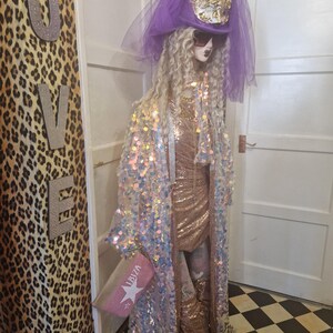 May include: A mannequin wearing a long, iridescent sequined robe with a pink and gold dress underneath. The mannequin is wearing a purple hat with a veil and sunglasses. The mannequin is holding a small pink clutch with the word "Ibiza" on it.