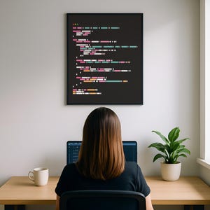 Puede incluir: Un cuadro enmarcado con líneas de código coloridas cuelga sobre un escritorio. Una persona está sentada en el escritorio, trabajando en una computadora portátil. Una taza blanca y una planta en maceta también están en el escritorio. La impresión de código tiene un fondo negro con líneas rosas, turquesas y blancas.