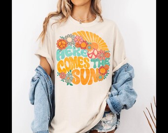 May include: A cream coloured t-shirt with a retro graphic design featuring a peace sign, flowers, and the text "Here Comes The Sun".