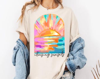 May include: Cream-colored t-shirt featuring a vibrant sunset design. The artwork depicts a colorful sunset over water, with the words "chasing sunsets" in a script font. The shirt has short sleeves and a relaxed fit.