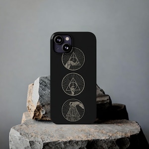 May include: Black phone case featuring three circular designs with hands and geometric shapes. Each design is within a triangle, with radiating lines. The phone case is resting on a gray stone surface.