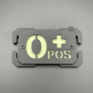 May include: A black metal tag with a white glow-in-the-dark O+ POS inscription. The tag has two holes on each side for attaching to a backpack or other gear.