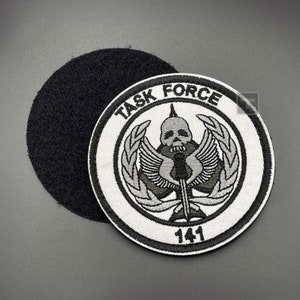 May include: White embroidered patch with a black skull and crossbones, wings, and laurel wreath. The text "TASK FORCE 141" is printed in black around the design.