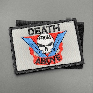 Pode incluir: Um remendo bordado branco com costuras pretas. O remendo apresenta uma caveira e ossos cruzados vermelhos e azuis com o texto "DEATH FROM ABOVE" em letras vermelhas e azuis.