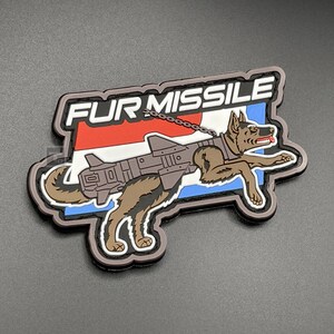 Pode incluir: Um patch com o texto "FUR MISSILE" acima de uma ilustração de um cão pastor alemão com um míssil preso. O patch tem um fundo vermelho, branco e azul, com uma borda preta. O cão é marrom e castanho claro.
