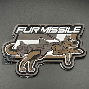 Puede incluir: Un parche bordado marrón y blanco con un pastor alemán que lleva un arnés y está atado a un cohete. El texto "FUR MISSILE" está por encima del perro.