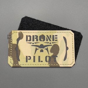 Peut inclure: Un patch de camouflage kaki et vert avec des coutures noires autour des bords. Le patch porte les mots "DRONE PILOT" en lettres noires avec une silhouette de drone entre les mots.