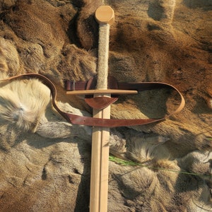 Puede incluir: Una espada de juguete de madera con una correa de cuero marrón y un mango de cuerda. La espada está sobre una superficie de piel marrón.