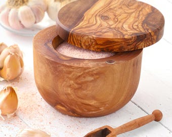 May include: A round wooden salt cellar with a matching lid, filled with pink Himalayan salt. A small wooden scoop rests beside the container. Several cloves of garlic are scattered nearby on a white surface, creating a kitchen scene.