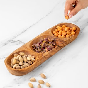 May include: A wooden serving tray with three separate compartments, each filled with different snacks. The compartments contain pistachios, small round orange snacks, and larger round orange snacks.