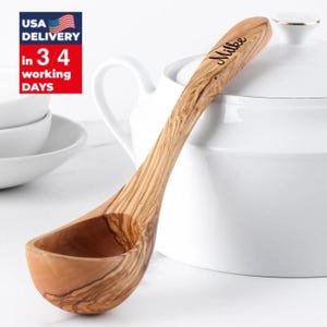 May include: A wooden olive wood ladle with a curved handle and a deep bowl. The handle is engraved with the name "Milke". The ladle is light brown with darker wood grain patterns. The ladle is next to a white ceramic pot.