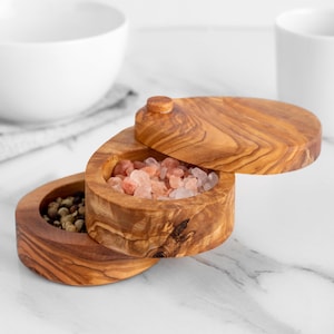 Könnte beinhalten: Ein Salz- und Pfefferstreuer-Set aus Holz mit Deckel. Das Set ist aus Olivenholz gefertigt und hat eine natürliche Oberfläche. Der Salzstreuer ist mit rosa Himalaya-Salz gefüllt und der Pfefferstreuer mit schwarzen Pfefferkörnern.
