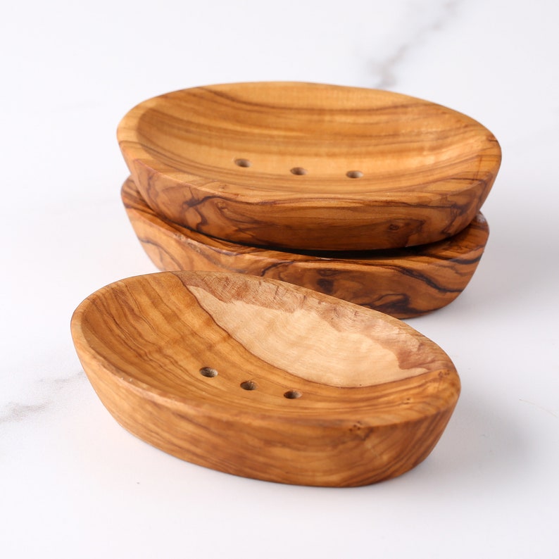 May include: Three wooden soap dishes made of olive wood. The dishes are oval shaped and have a natural wood finish. The dishes are stacked on top of each other.