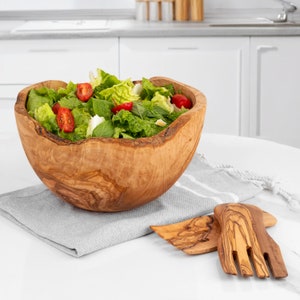 May include: A wooden salad bowl with a natural, rustic finish, filled with a green salad with cherry tomatoes. Two wooden salad servers are on a white surface next to the bowl.