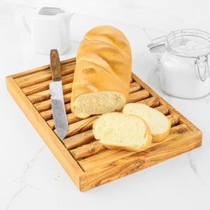 May include: A rectangular wooden bread board with a slatted surface, featuring a loaf of golden-brown bread, two sliced pieces, and a serrated knife with a wooden handle resting on the board, alongside a white pitcher and a glass jar.