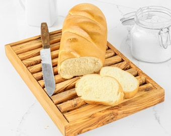 Puede incluir: Una tabla de pan rectangular de madera con superficie de listones, que presenta una hogaza de pan dorado, dos rebanadas y un cuchillo dentado con mango de madera sobre la tabla, junto a una jarra blanca y un frasco de vidrio.