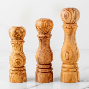 以下が含まれることがあります： さまざまなサイズの木製ソルト＆ペッパーグラインダー3つ。グラインダーは、天然の木目模様の明るい茶色の木材で作られています。各グラインダーは、丸みを帯びた上部と、ベース近くに装飾リングが付いた円筒形のボディを備えています。