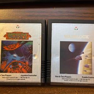 以下が含まれることがあります： 2つのビンテージビデオゲームカートリッジ。「バミューダトライアングル」カートリッジは、コックピットと水中シーンのカラフルなグラフィックが特徴です。「ウォーロック」カートリッジは、惑星と宇宙船のある宇宙シーンを示しています。どちらも1人または2人用のゲームです。