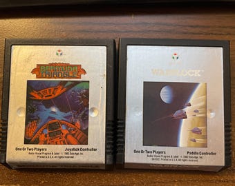 Könnte beinhalten: Zwei Vintage-Videospielkassetten. Die "Bermuda Triangle"-Kassette zeigt eine farbenfrohe Grafik eines Cockpits und einer Unterwasserszene. Die "Warlock"-Kassette zeigt eine Weltraumszene mit Planeten und Schiffen. Beide sind für ein oder zwei Spieler.