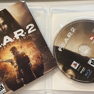 Może przedstawiać: Etui na grę wideo PlayStation 3 z grą "F.E.A.R. 2: Project Origin". Etui jest białe z czarną płytą w środku. Okładka gry przedstawia kobietę z długimi ciemnymi włosami i mężczyznę trzymającego broń.