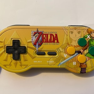 Puede incluir: Un mando de videojuegos amarillo y negro con ilustraciones de "The Legend of Zelda: A Link to the Past". El mando tiene una cruceta, botones y el logotipo del juego. Está conectado a un cable negro.