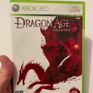 Może przedstawiać: Pudełko gry Xbox 360 do gry Dragon Age: Origins. Okładka przedstawia czerwonego smoka, wojownika i kobietę z czerwonymi włosami. Gra jest oceniona przez ESRB jako M dla dorosłych.