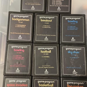 May include: A collection of 12 Atari game cartridges, each with a black plastic case and a gold label. The labels feature the game title, game program logo, game number, and instructions for use. The games include Championship Soccer, Video Olympics, Backgammon, Breakout, Bowling, Football, Sky Diver, Space Invaders, Basketball, and Flag Capture.