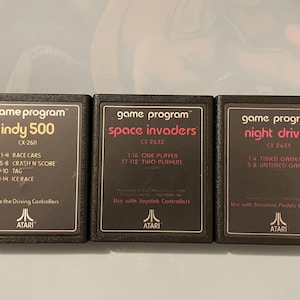 Puede incluir: Tres cartuchos de juego Atari para la consola Atari 2600. Los cartuchos son negros con texto blanco y un logotipo de Atari rojo. Los títulos de los juegos son "Indy 500", "Space Invaders" y "Night Driver".