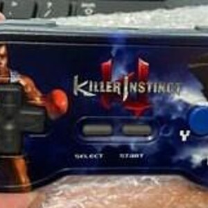 Puede incluir: Un mando de videojuegos negro y azul de Killer Instinct con el logotipo del juego y gráficos de personajes. El mando tiene una cruceta, botones de selección e inicio, y cuatro botones de acción etiquetados X, A, Y y B. El mando lo sostiene una persona.