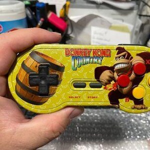 Puede incluir: Un mando de videojuego amarillo de Donkey Kong Country con un barril y el personaje Donkey Kong. El mando tiene botones rojos, amarillos y negros. El mando está en la mano de una persona.