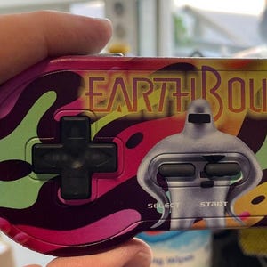 Pode incluir: Um controle de videogame colorido com o texto "EARTH BOUND" em laranja. O controle tem um direcional preto, botões laranja e verdes e um gráfico de robô prateado. O design apresenta redemoinhos de rosa, verde e amarelo.