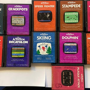 Puede incluir: Una colección de cartuchos de videojuegos Activision vintage, con títulos como "Star Master", "Crackpots" y "Spider Fighter". Cada cartucho tiene una combinación de colores única y gráficos de juego en la parte delantera, mostrando la estética clásica de los juegos.