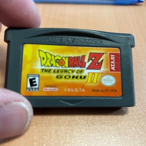 May include: A Game Boy Advance cartridge for Dragon Ball Z: The Legacy of Goku II. The label features the game title in yellow and red, with the Atari logo. The cartridge is black and held by a person's fingers.
