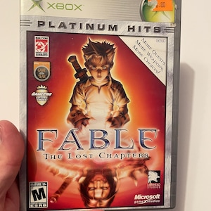 May include: Xbox game case for Fable: The Lost Chapters. The case is silver with a red background and features a young boy holding a sword. The game is rated M for Mature by the ESRB.