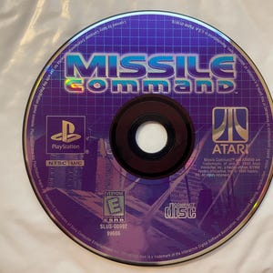 May include: A purple PlayStation game disc for Missile Command. The disc features the game's title in large, blue and white letters, along with the PlayStation and Atari logos. The disc is rated "Everyone" by the ESRB.