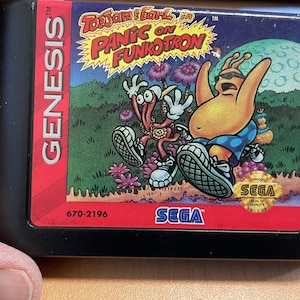 May include: A black Sega Genesis game cartridge for "ToeJam & Earl in Panic on Funkotron." The label displays vibrant cartoon graphics of two alien characters. The cartridge features the "Genesis" logo and the Sega logo.