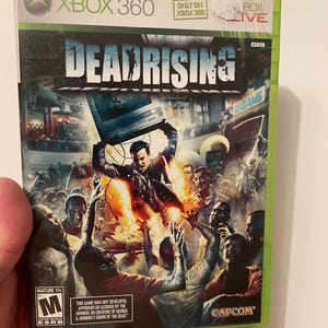 以下が含まれることがあります： Xbox 360用ゲームケース、デッドライジング。17歳以上対象のゲームで、赤、オレンジ、黄色のカラーリング。カバーアートは、テレビを頭上に掲げた男性が、ゾンビに囲まれている様子を描いている。