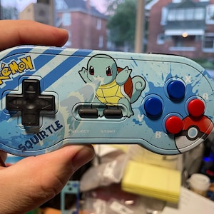 Könnte beinhalten: Ein hellblauer Videospiel-Controller mit dem Pokémon-Charakter Schiggy. Der Controller hat ein schwarzes Steuerkreuz, schwarze Tasten und blaue und rote Aktionstasten. Das Wort "Pokémon" ist oben und "Schiggy" unten aufgedruckt.