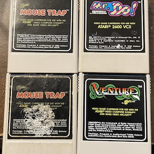 Op de afbeelding: Vier vintage Atari-videospelcartridges. De cartridges zijn wit met zwarte tekst en afbeeldingen. De spellen zijn Mouse Trap, Mr. Do!, Mouse Trap en Venture.