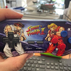 Op de afbeelding: Een handheld videogamecontroller met een Street Fighter II-afbeelding. De controller heeft een zwart kruis, selectieknoppen en rode en blauwe actieknoppen. De afbeelding toont twee personages in vechthoudingen.