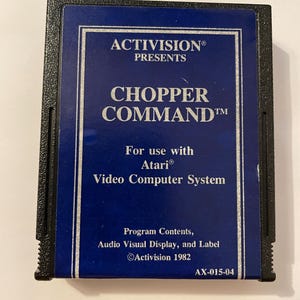 May include: A vintage Atari game cartridge for "Chopper Command" by Activision. The cartridge has a dark blue label with white text, including the game title and "Activision Presents." The cartridge is black.